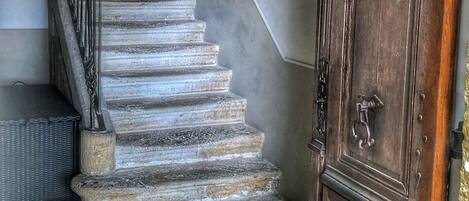 A staircase with stone steps leading to a landing, a wooden door to the right, and framed pictures on the walls.