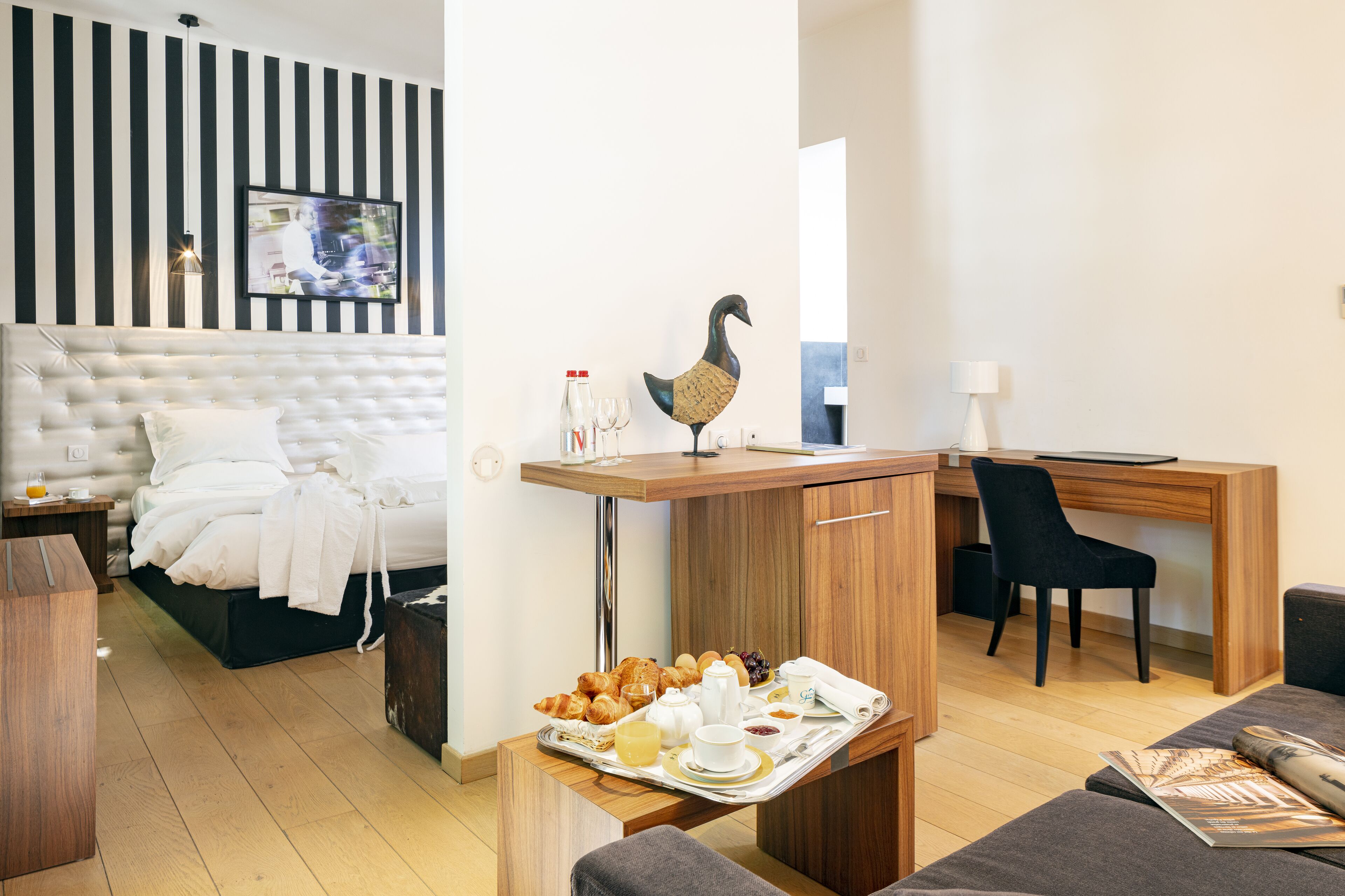 Habitación de hotel moderna con una cama grande, un escritorio de madera, una silla y una bandeja de desayuno con cruasanes y bebidas.