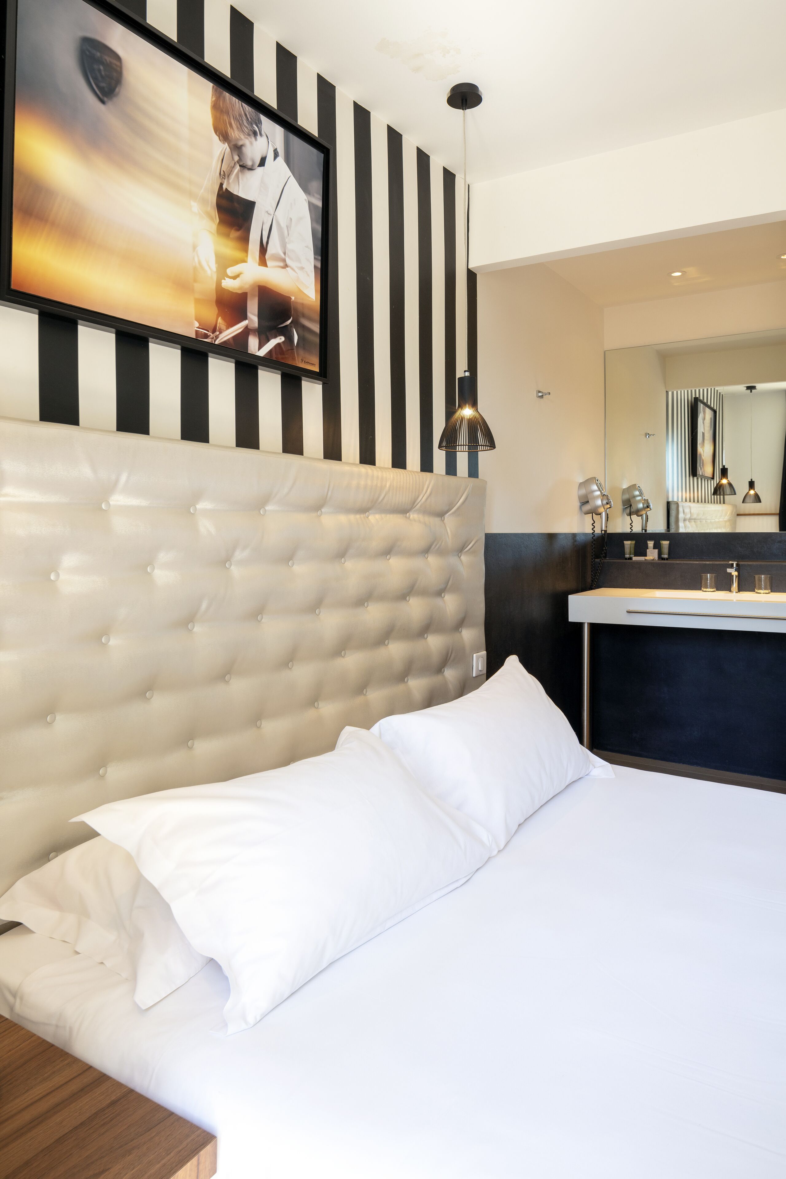 Ein Hotelzimmer mit einer gesteppten Kopfteil, einem großen Bett mit weißen Leinentüchern, einem modernen Badezimmer mit Waschbecken und Spiegel sowie einem gerahmten Bild an der Wand.