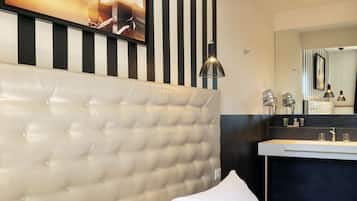 Habitación de hotel con cabecera capitonada, cama grande con ropa blanca, baño moderno con lavamanos y espejo, y un cuadro enmarcado en la pared.