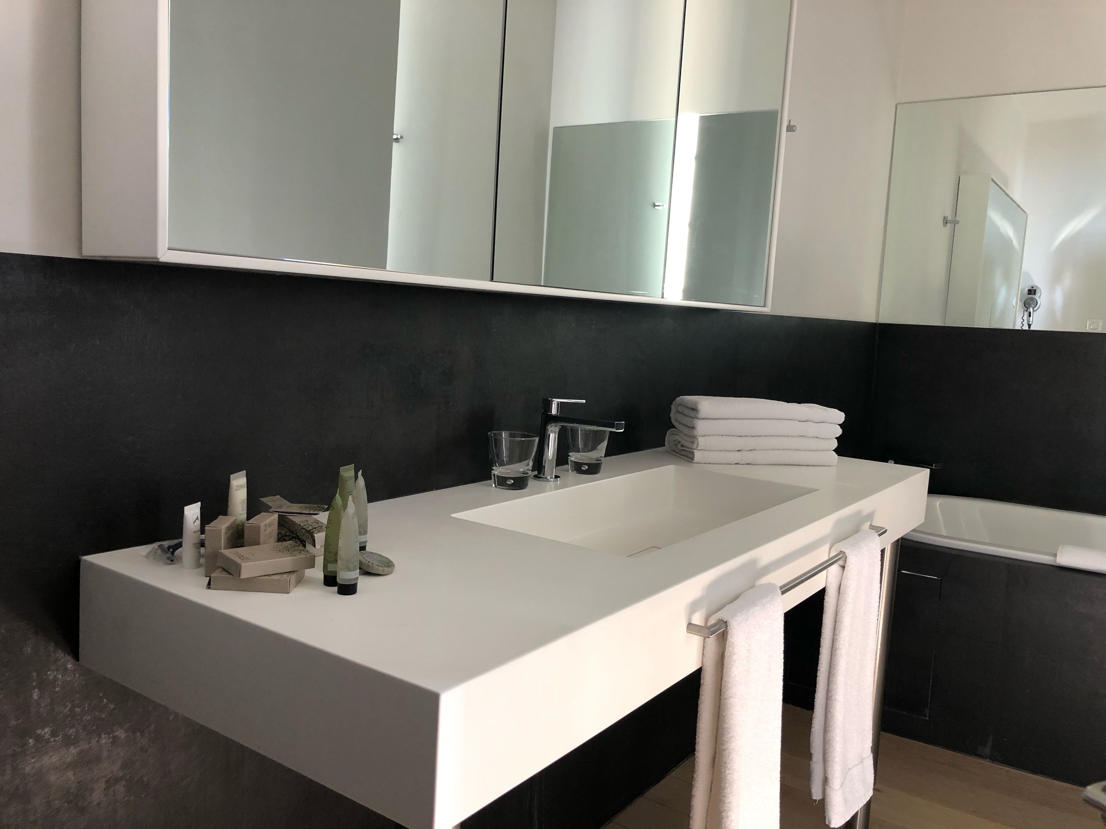 A modern bathroom with a white vanity, a large mirror, a bathtub, and a dark wall.