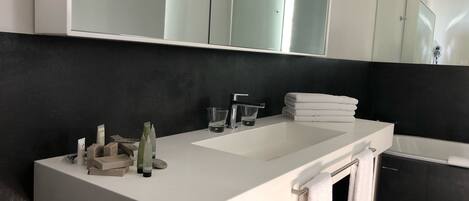 A modern bathroom with a white vanity, a large mirror, a bathtub, and a dark wall.