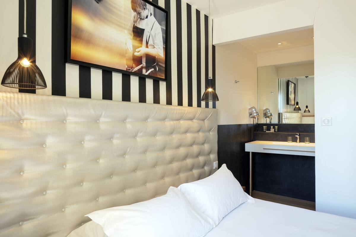 Habitación de hotel moderna con cabecera capitonada, un televisor de pantalla plana grande, pared a rayas blancas y negras, tocador oscuro con espejo e iluminación colgante.