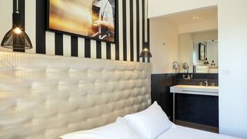 A modern hotel room with a tufted headboard, a large flat-screen TV, a black and white striped wall, a dark-toned vanity with a mirror, and pendant lighting.