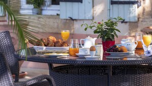 Une table de petit-déjeuner installée à l’extérieur, avec une chaise en osier, un panier de croissants, une plante en pot et un verre de jus d’orange.