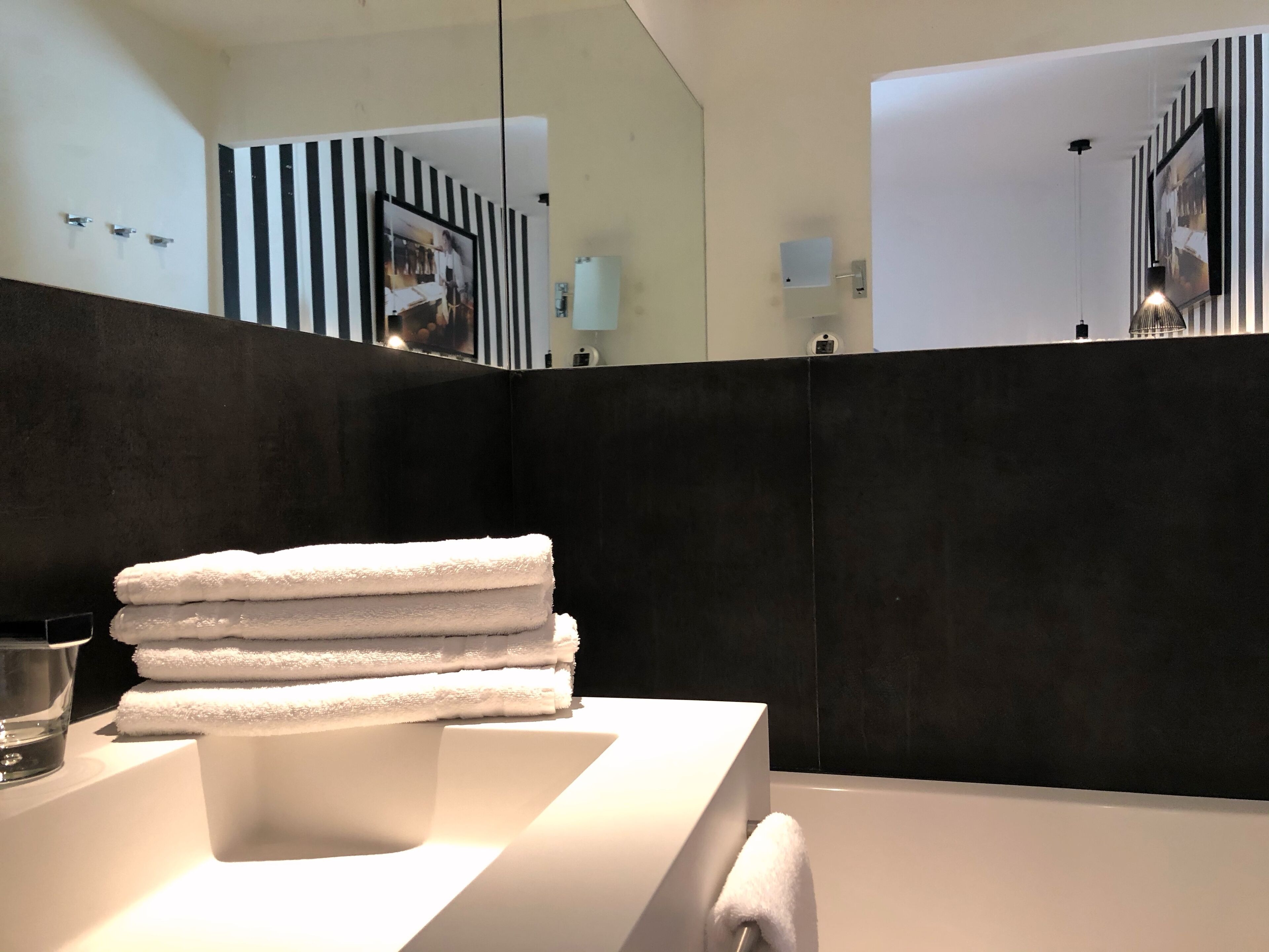 Ein modernes Badezimmer mit einem weißen Waschbecken, einem Stapel weißer Handtücher und einem großen Spiegel, der den Raum widerspiegelt.