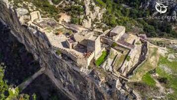 Vista aérea de una fortificación histórica con varios niveles y muros, rodeada de vegetación y terreno rocoso.