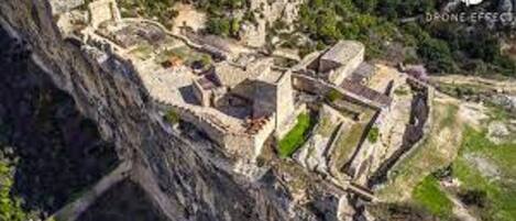 Vista aérea de una fortificación histórica con varios niveles y muros, rodeada de vegetación y terreno rocoso.