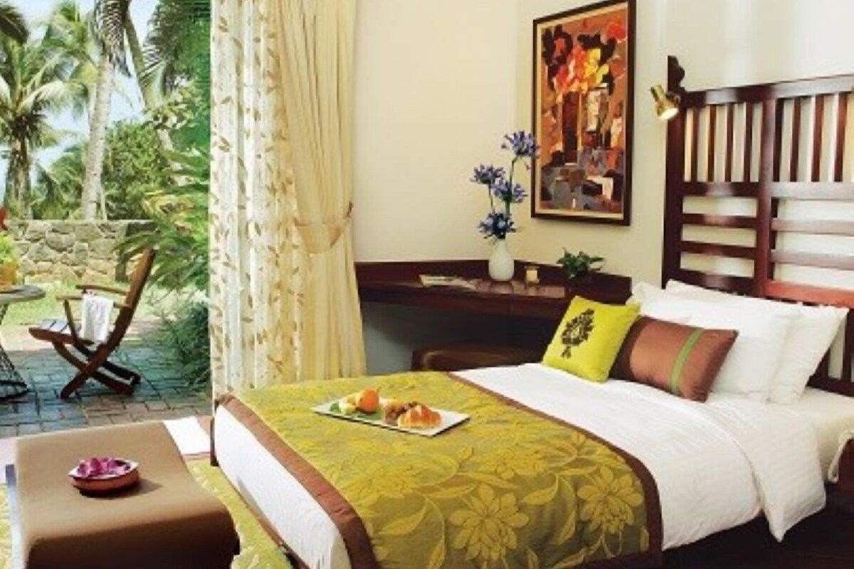 A hotel room with a bed, a desk, a chair, a window with a view of palm trees, and a painting on the wall.