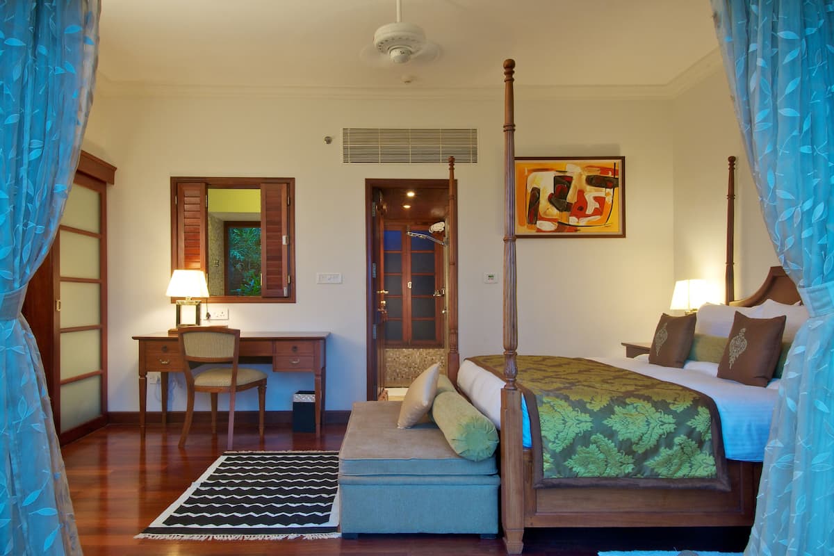 A bedroom with a four-poster bed, a desk with a chair, a bedside table, and a large window.