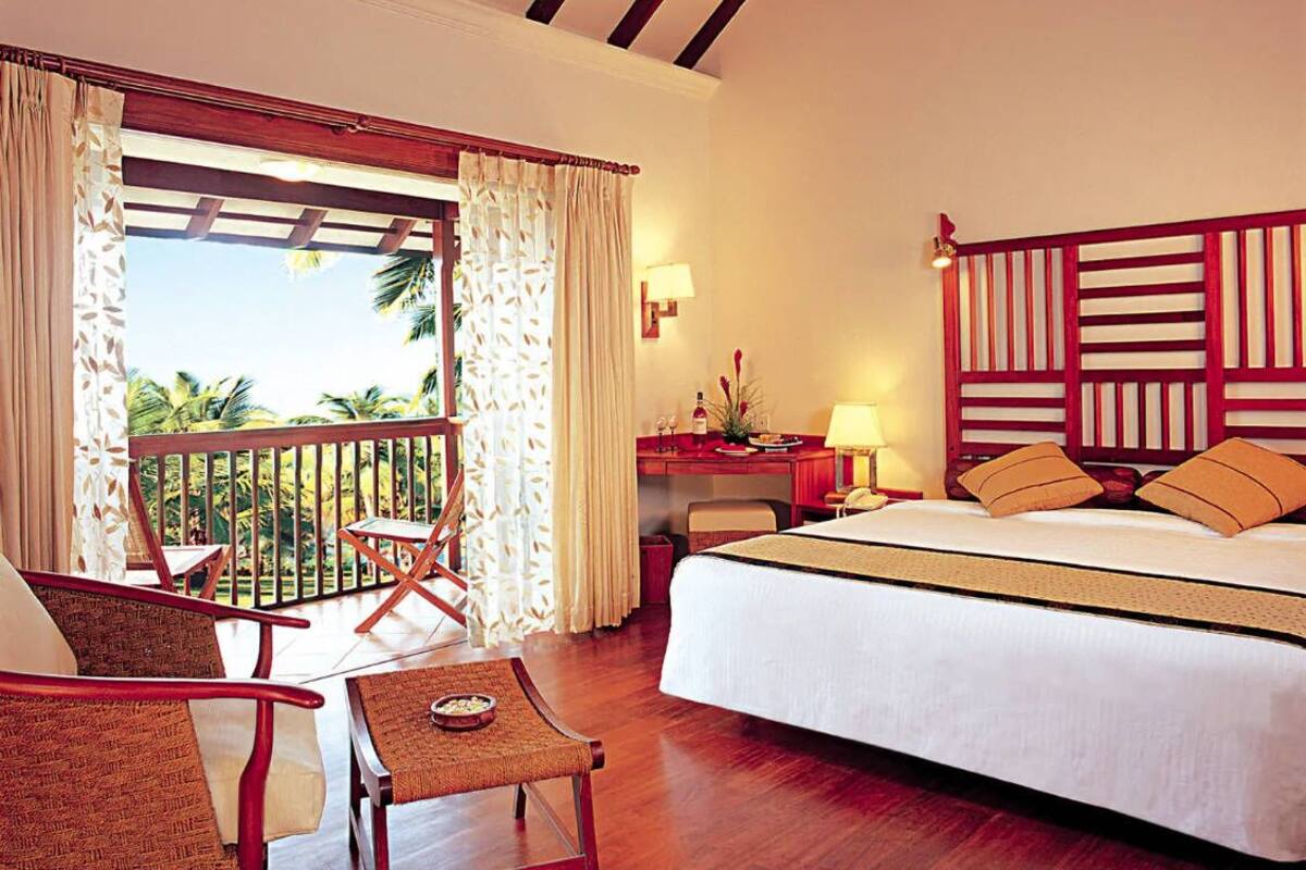 A hotel room with a bed, a desk, a chair, a small table, and a balcony with a view of palm trees.