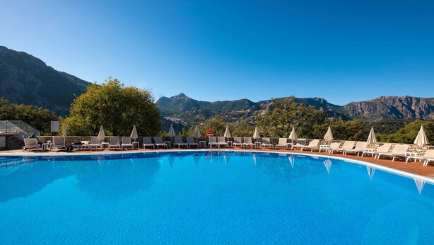 A pool with lounge chairs and umbrellas, surrounded by trees and mountains.