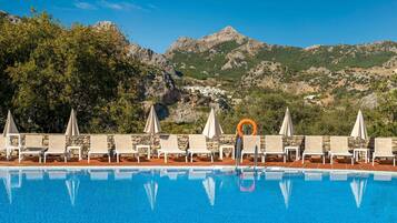 Piscina con sillones de descanso y sombrillas, rodeada por un muro de piedra y vistas a montañas y un pueblo.