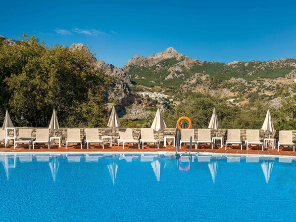 Piscina con sillones de descanso y sombrillas, rodeada por un muro de piedra y vistas a montañas y un pueblo.