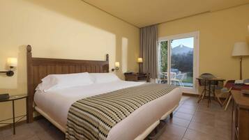 A hotel room with a large bed, a desk with a chair, a small table with chairs, and a view of the outdoors through a sliding door.