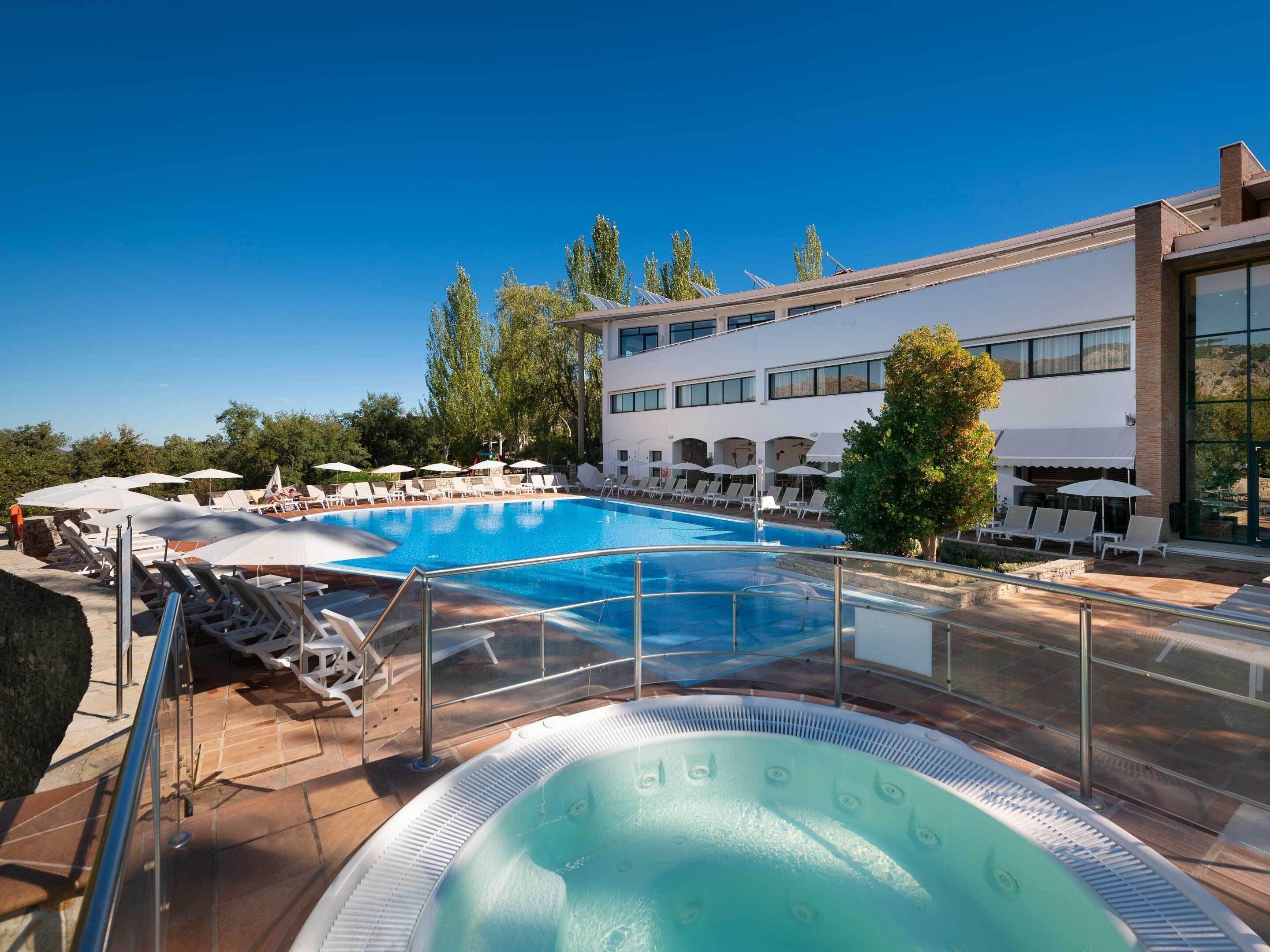 Una piscina con lettini e ombrelloni, circondata da un edificio con ampie finestre e una zona patio.