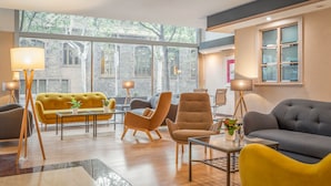 A modern living room with a yellow sofa, grey armchairs, and a grey sofa. There are two floor lamps, a coffee table, and a view of a stone building through large windows. - Sunotel Aston (Barcelona)