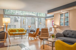 A modern living room with a yellow sofa, grey armchairs, and a grey sofa. There are two floor lamps, a coffee table, and a view of a stone building through large windows.
