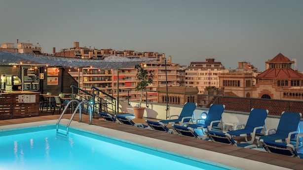 A rooftop pool with blue lounge chairs, a bar area, and a view of the cityscape. - Sunotel Aston (Barcelona)