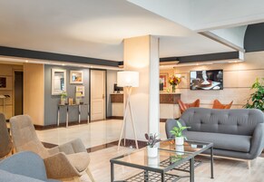A modern hotel lobby with a seating area, a coffee station, and a television displaying a meal.