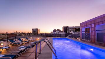 Una piscina en la azotea con sillones de descanso y vistas a la ciudad al atardecer.