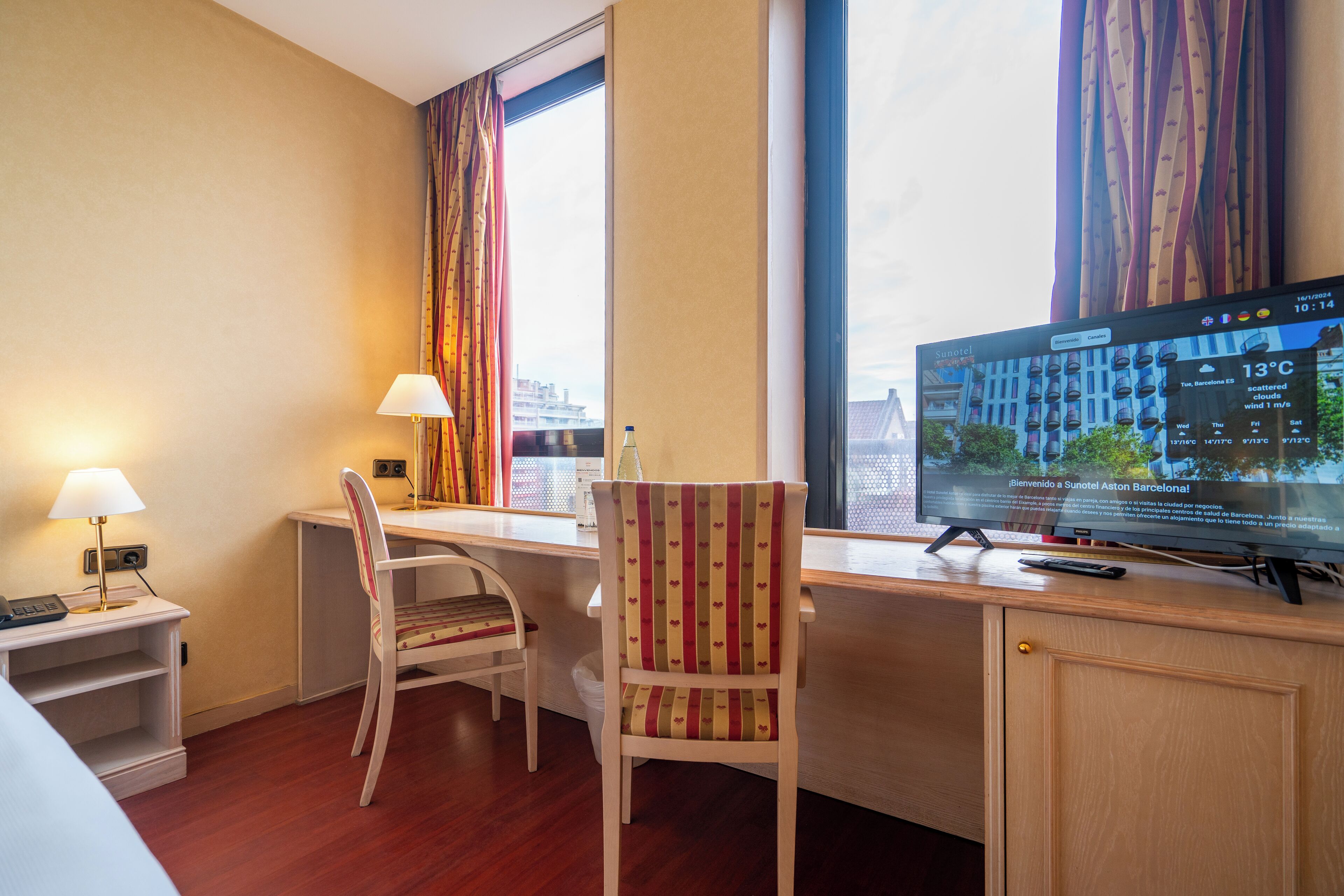 Een hotelkamer met een bureau, twee stoelen, een televisie en een raam met gordijnen.