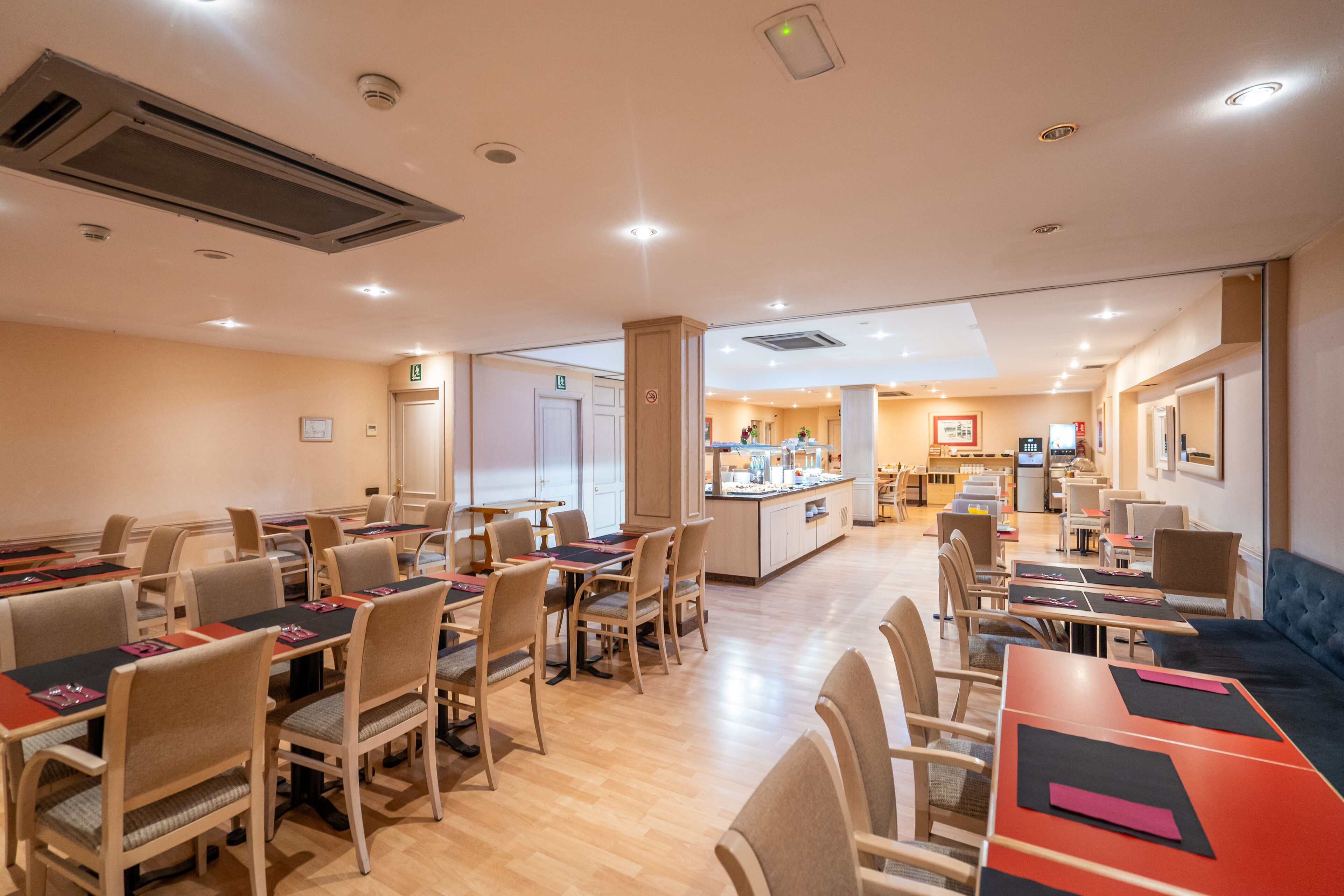 Un comedor de hotel con varias mesas y sillas, una isla central con cocina, y un espacio de descanso con sofá.