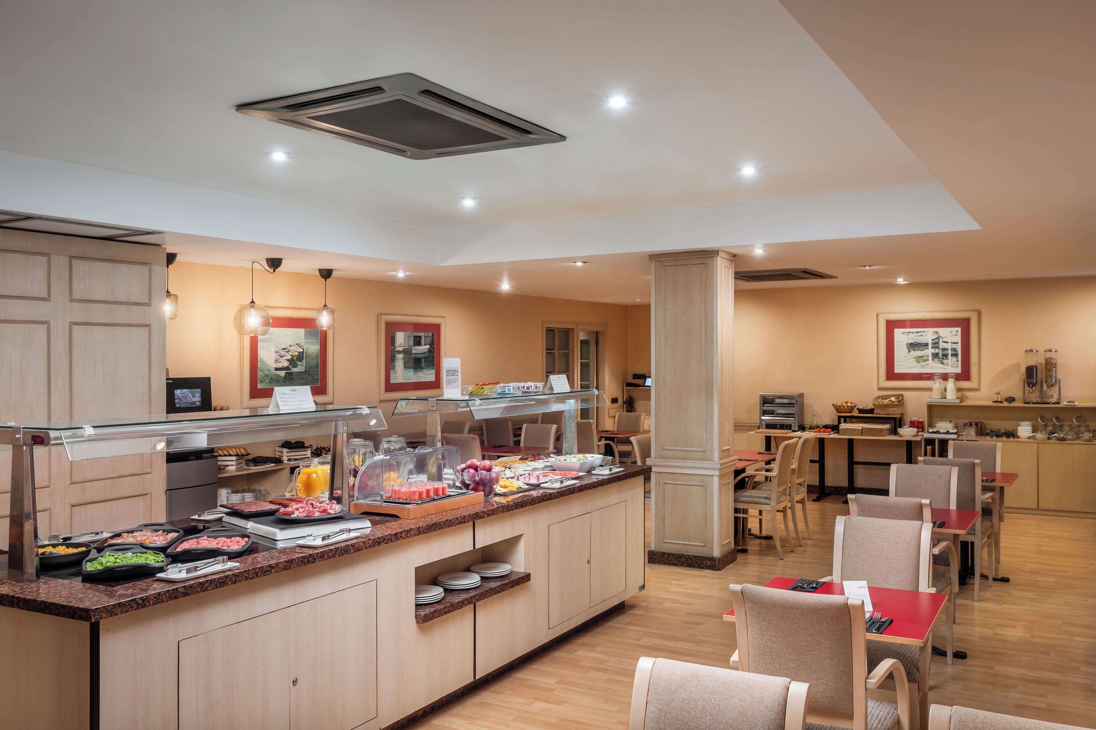 Comedor de hotel con una barra de buffet, disposición de mesas y sillas, y una zona de cocina al fondo.