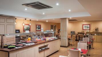 Comedor de hotel con una barra de buffet, disposición de mesas y sillas, y una zona de cocina al fondo.
