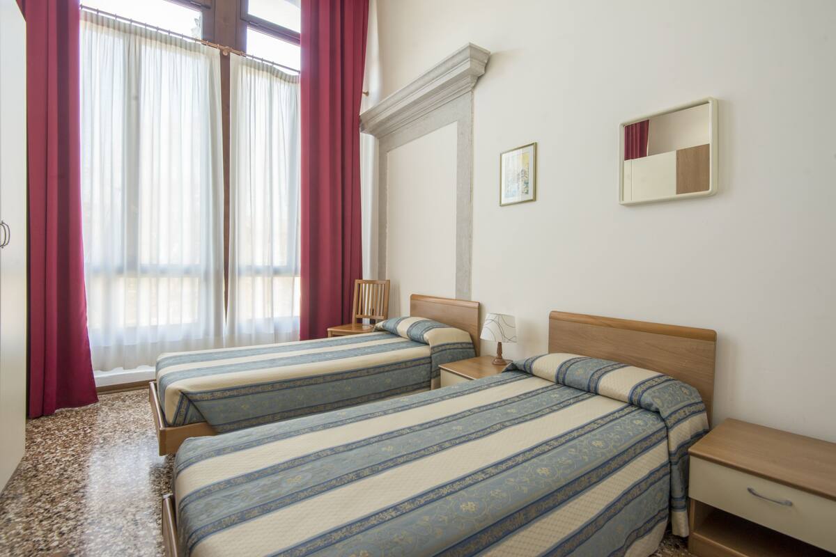 A hotel room with two beds, a nightstand, a bedside table, a mirror, and a window with curtains.