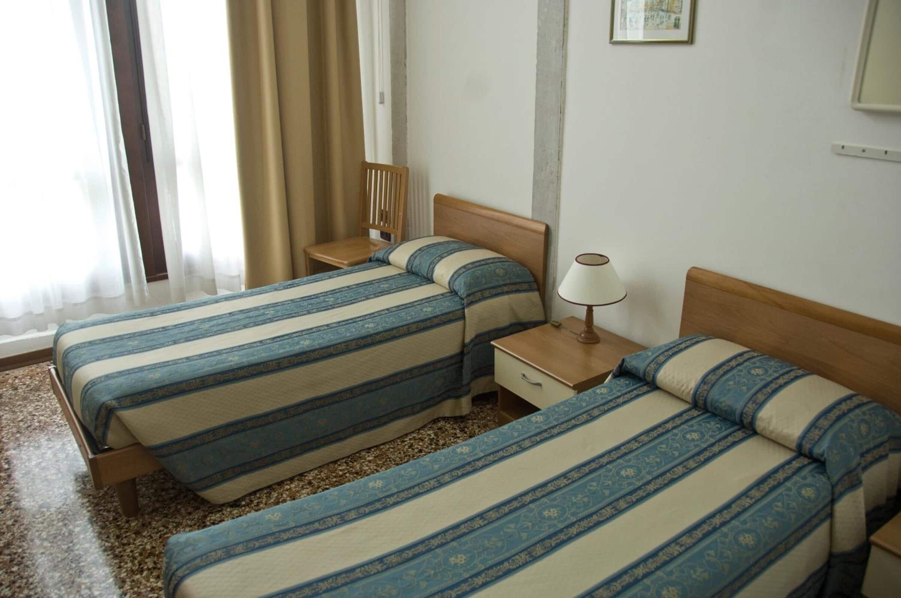 A hotel room with two beds, a nightstand, a lamp, and a chair.