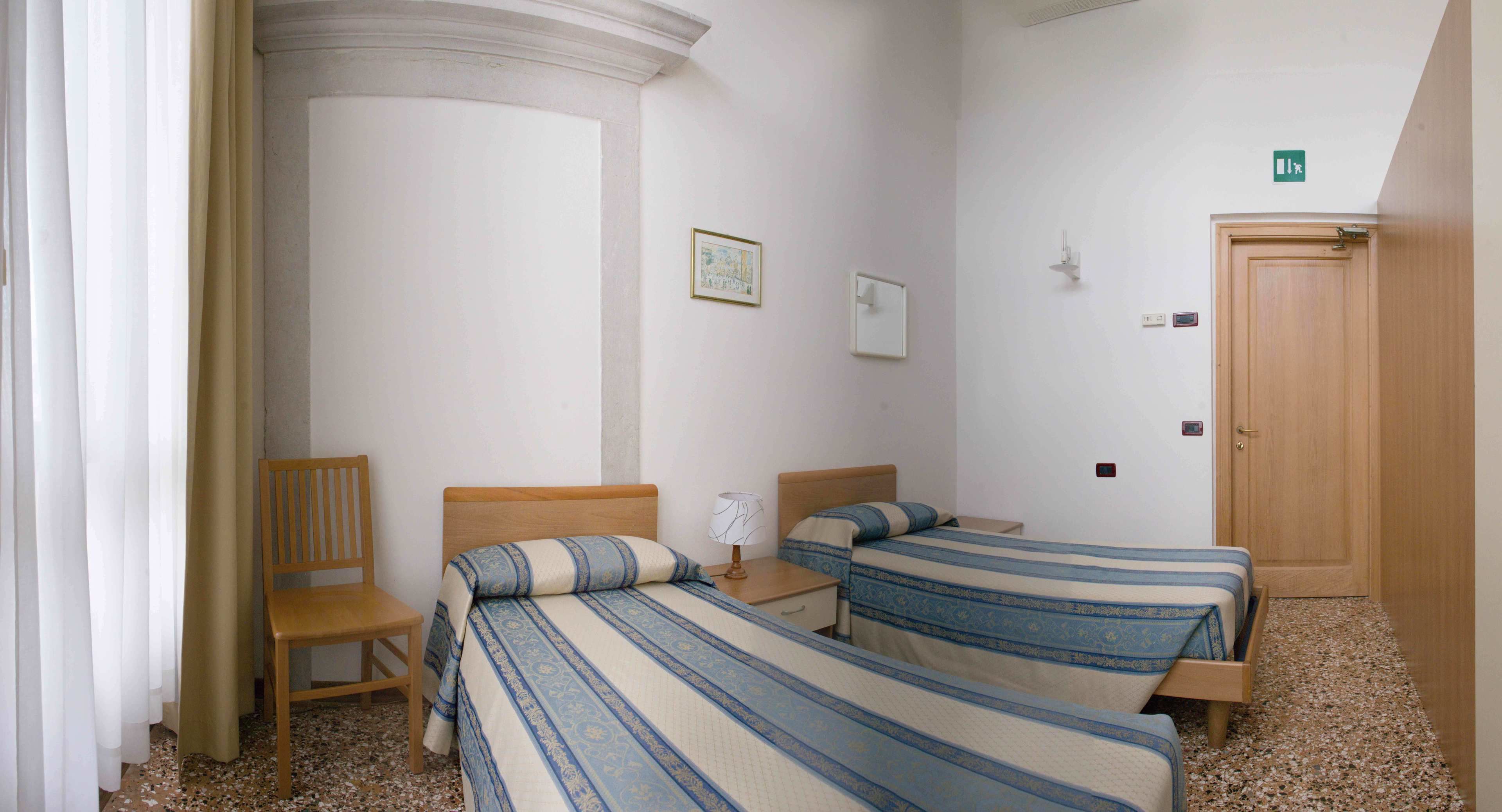 Habitación de hotel con dos camas individuales, una silla de madera, una mesita de noche con lámpara y una puerta con letrero de salida de emergencia.