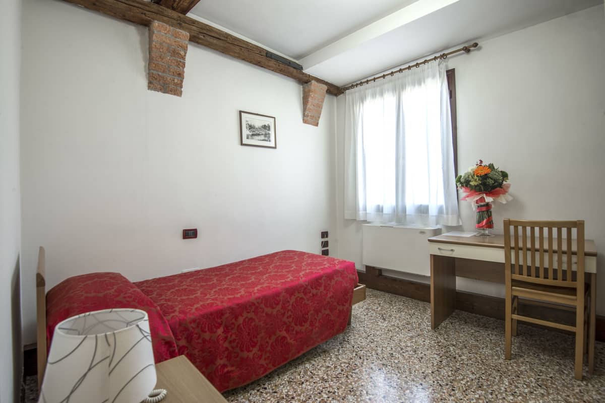 A small, simple room with a bed, a desk, a chair, and a window with curtains.