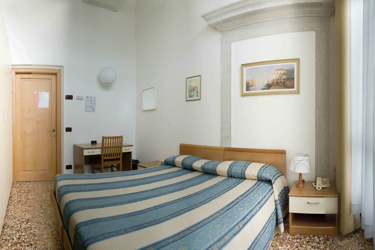 A hotel room with a bed, a desk, a chair, a nightstand with a lamp, and a painting on the wall.