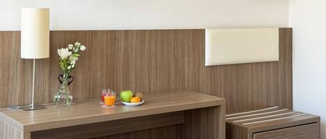 Una habitación de hotel moderna con un escritorio de madera, una mesita de noche, una lámpara, una planta pequeña y un plato de fruta.