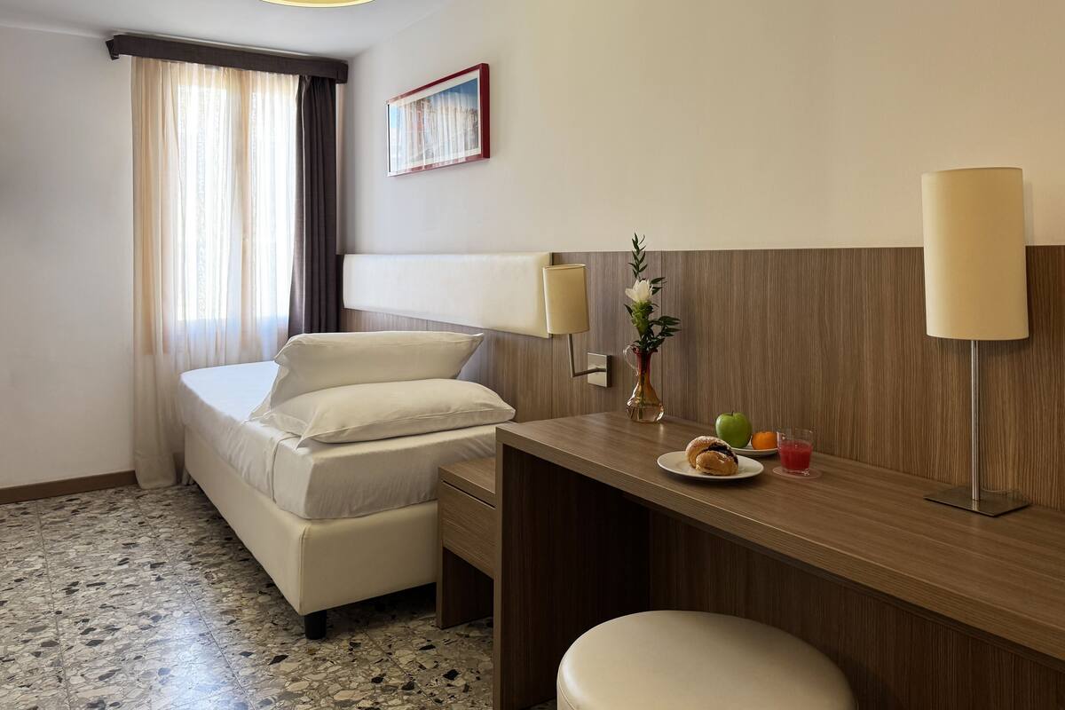 A hotel room with a bed, a desk, a chair, a lamp, a vase with flowers, and a fruit plate.