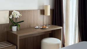 Quarto de hotel com escrivaninha de madeira, um vaso branco com flores brancas, uma luminária, um prato com pastelaria e um banquinho branco.