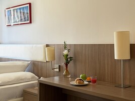 A hotel room with a bed, bedside table, lamp, vase with flowers, plate with food, and a glass of drink.