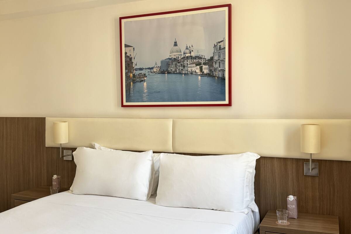 A hotel room with a bed, two bedside lamps, a nightstand with a glass and a water bottle, and a framed picture of a canal scene on the wall.