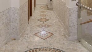 Um corredor com piso de mármore, uma porta de vidro à direita e um tapete estampado no final.