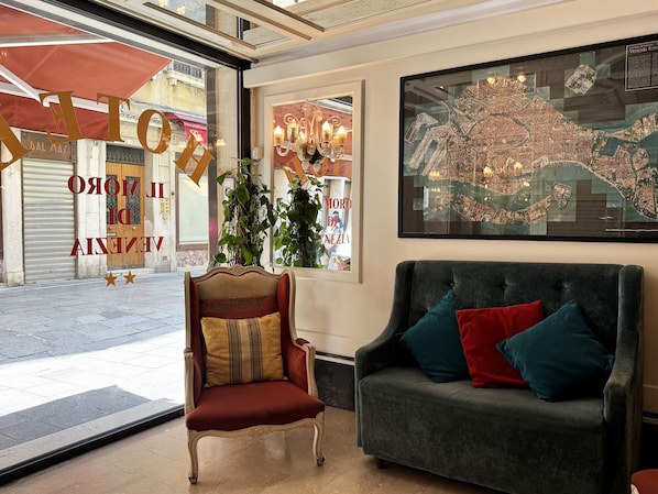 A lobby with a red velvet armchair, a grey sofa with colorful cushions, a large framed map, and a window with a view of the street.