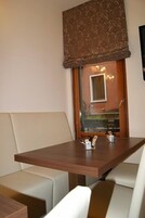 Una habitación moderna con una mesa de madera y dos sillas blancas acolchadas. Se ve una ventana grande con una persiana estampada, y un espejo pequeño cuelga en la pared.