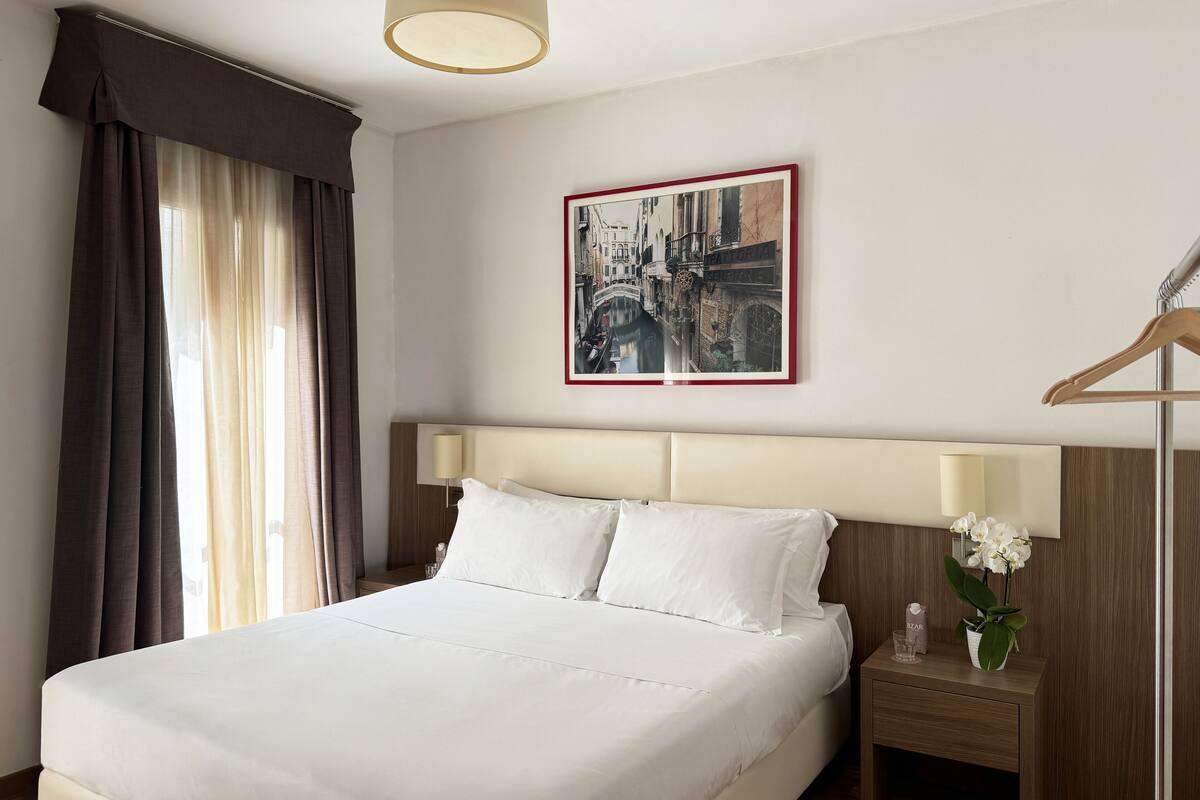 A hotel room with a large bed, a nightstand with a lamp, a framed picture on the wall, and a wooden hanger.