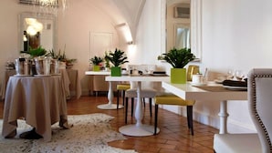 Un coin repas avec des tables et des chaises blanches, ainsi qu’un lustre. Des plantes en pot sont disposées sur les tables et un tapis blanc recouvre le sol.