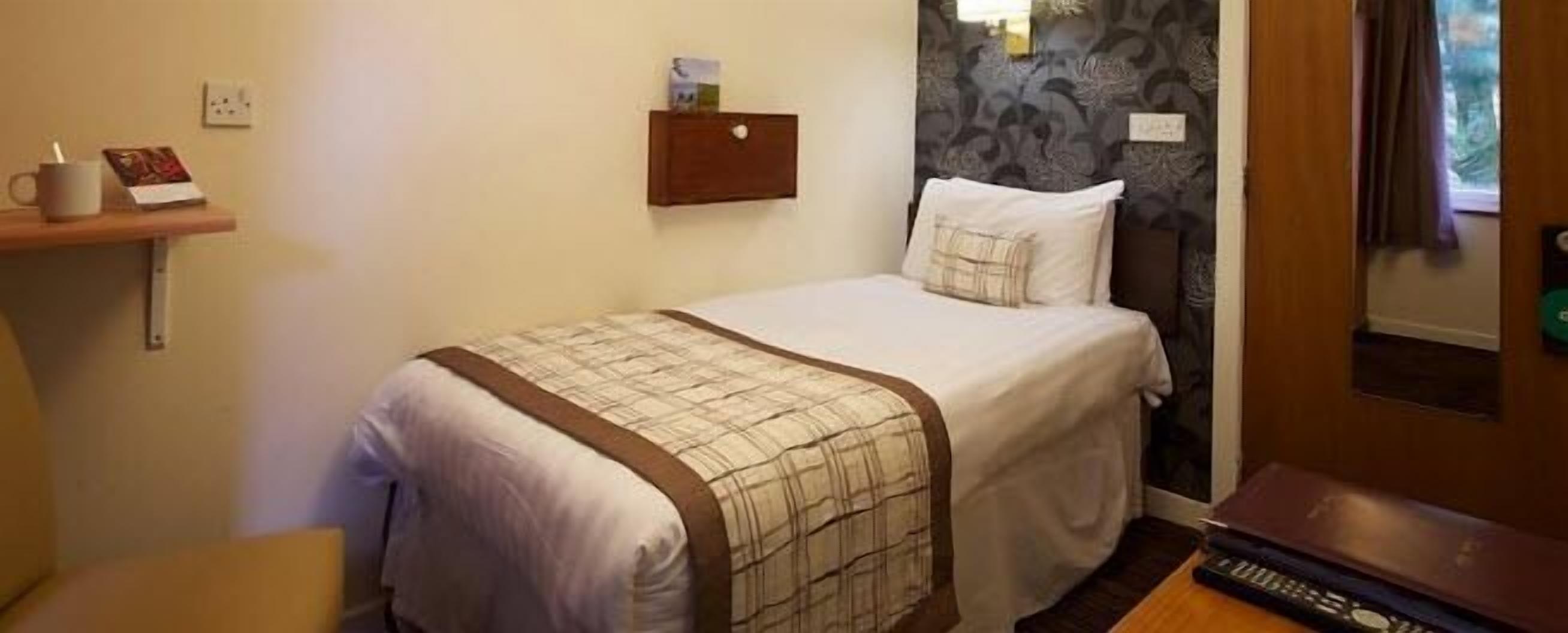 A neatly made bed with a plaid bedspread, a bedside table with a book, and a wall-mounted shelf with a mug and a card.
