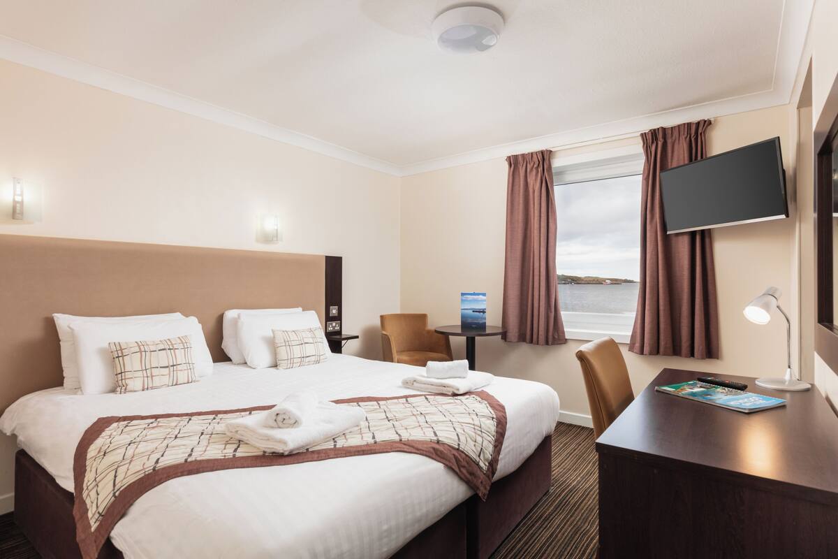 A hotel room with a large bed, a desk, a chair, a TV, and a window with curtains.