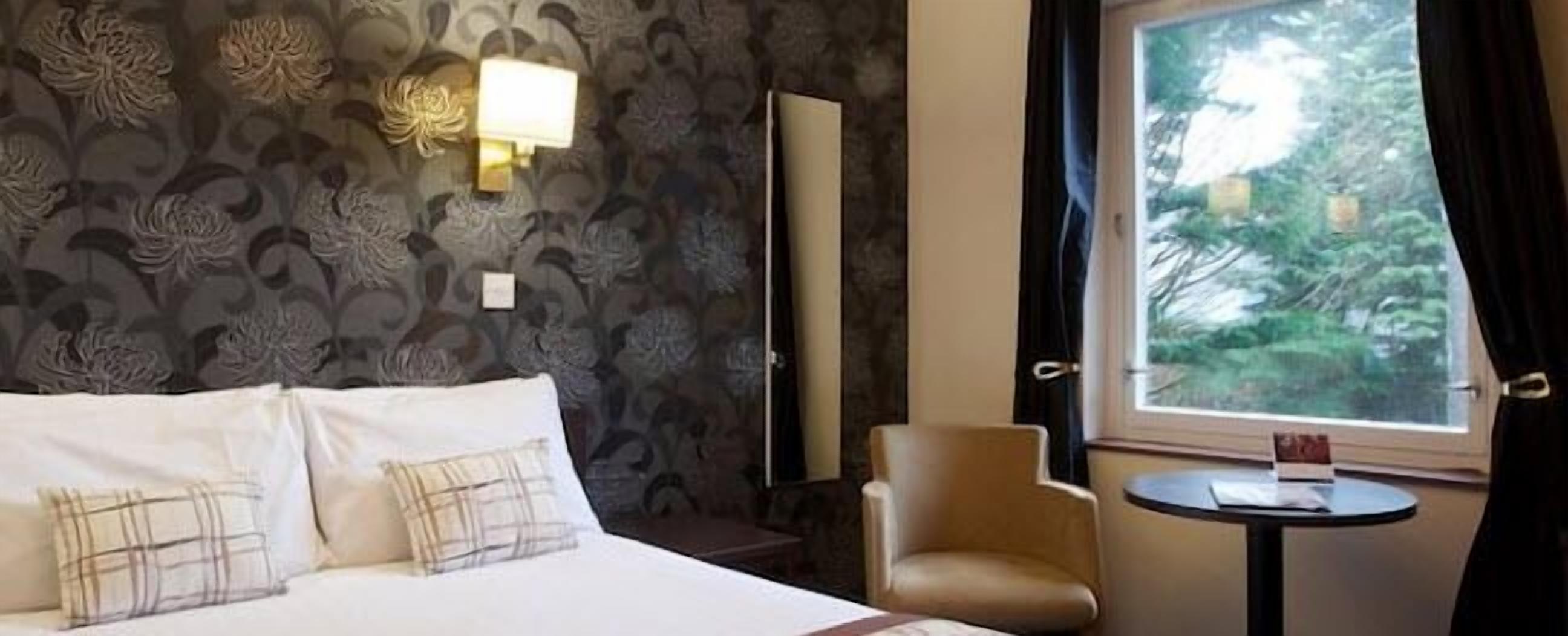 A hotel room with a bed, a chair, a small table, and a window with a view of trees.