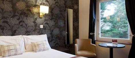 Ein Hotelzimmer mit Bett, Sessel, kleinem Tisch und Fenster mit Blick auf Bäume.