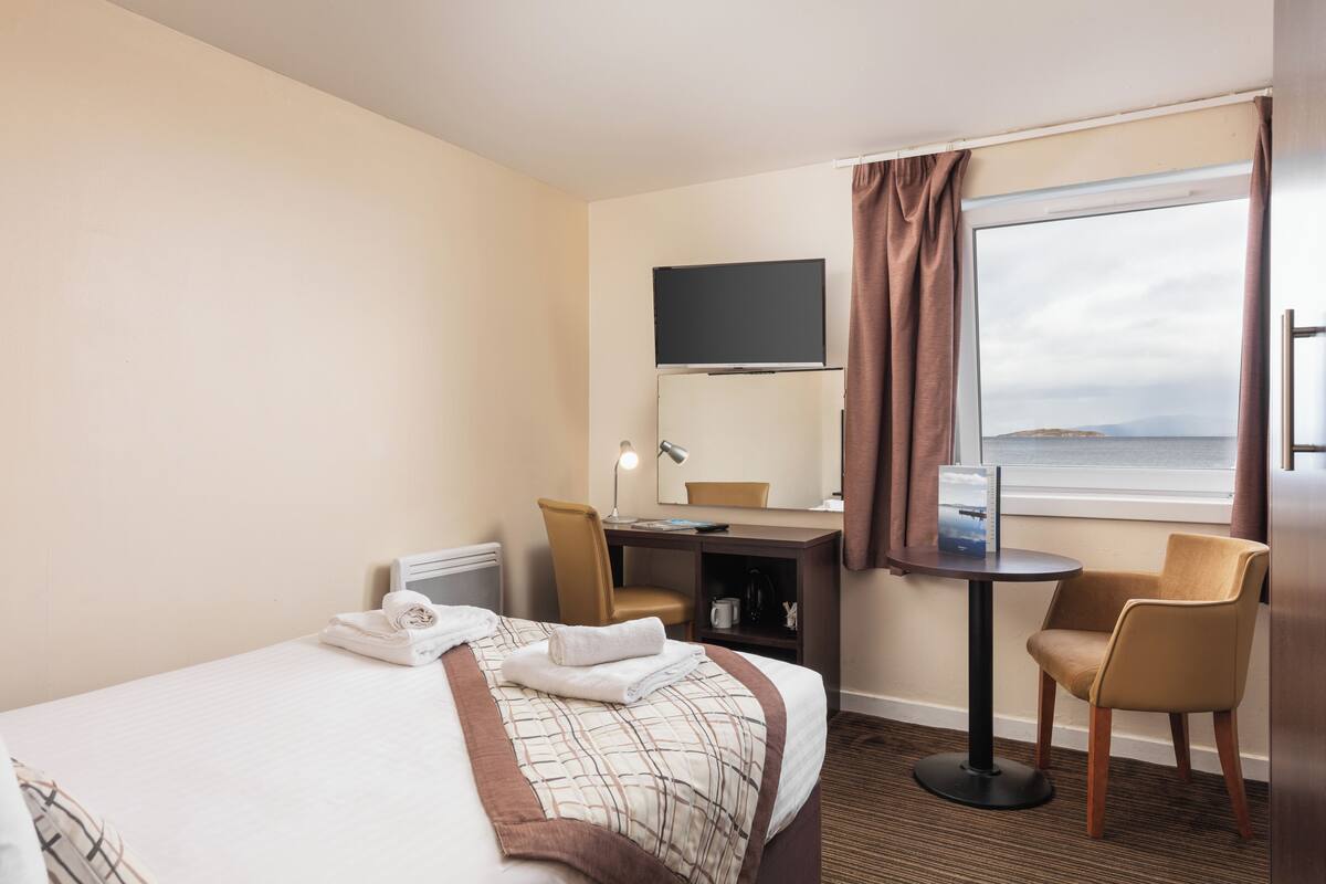 A hotel room with a bed, a desk, a chair, a TV, and a window with a view.