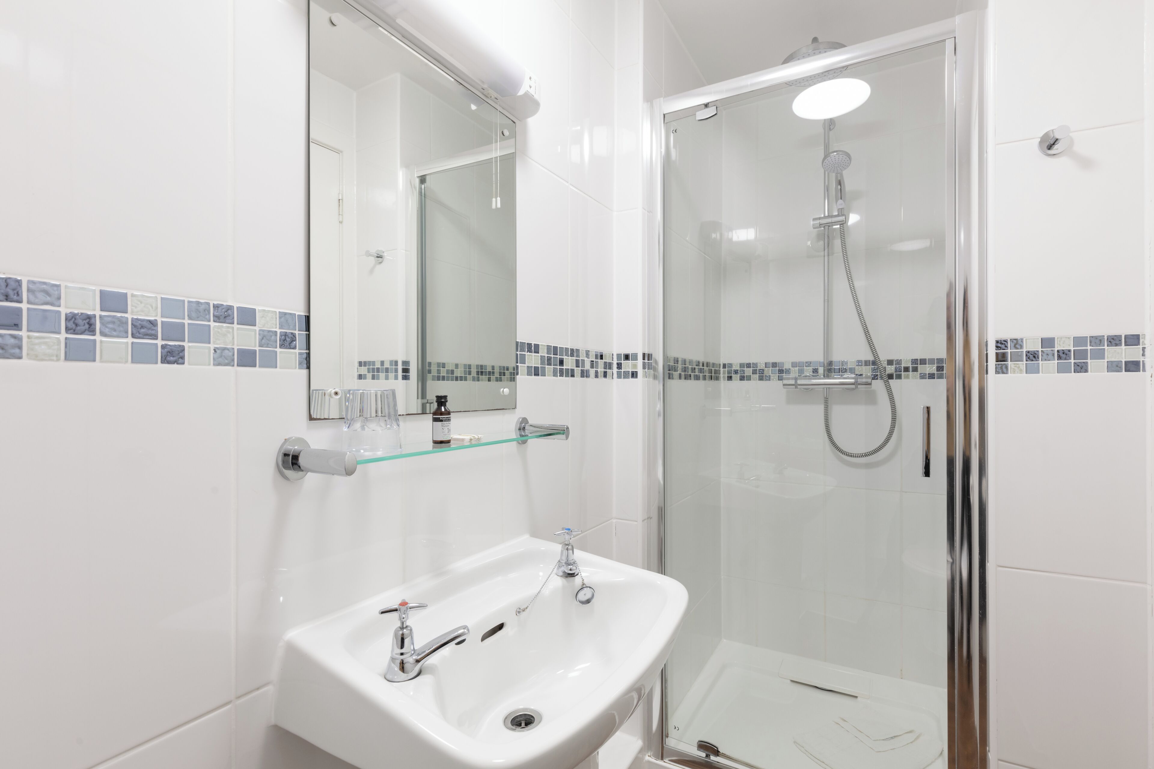 Un baño moderno con ducha envuelta en vidrio, un lavabo blanco con dos llaves y un estante de vidrio sobre el lavabo.